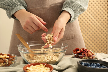 Making granola. Woman putting oat flakes into bowl at table in kitchen, closeup
