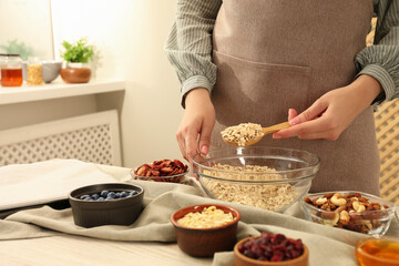 Making granola. Woman putting oat flakes into bowl at table in kitchen, closeup