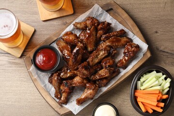 Tasty chicken wings, glasses of beer, sauces and vegetable sticks on wooden table, flat lay....