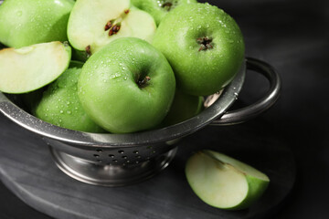 Ripe green apples with water drops and colander on black table, closeup