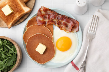 Tasty pancakes served with fried egg and bacon on white tiled table, flat lay