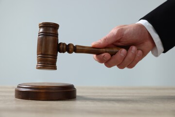 Judge with gavel sitting at wooden table against light grey background, closeup