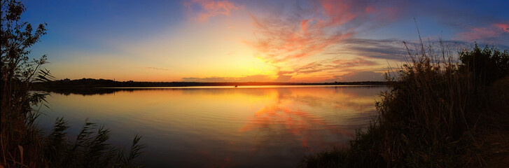 Stunning sunset on the shore of a lake with reeds in the foreground. panorama