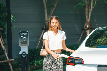 Young woman travel with EV electric car charging in green sustainable city outdoor garden in...