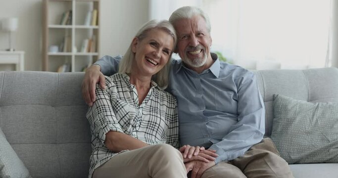 Attractive older retired husband and wife hugging with heads touch on cozy home couch, looking at camera, getting happy, positive, cheerful, laughing, holding hands. Family portrait shot