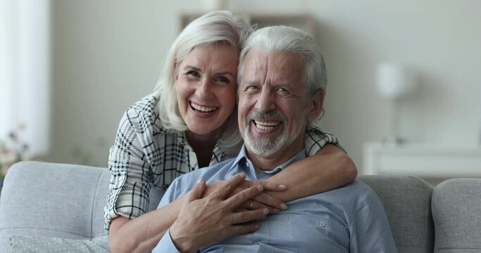 Happy positive senior married couple in love hugging on couch, posing for home portrait, looking at camera, laughing, smiling with healthy white teeth, enjoying romantic relationship, retirement