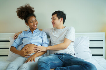 American - African black ethnicity woman and her Asian husband relaxing together on the bed in bedroom and a man tenderly touching on his wife belly. Husband taking care of his pregnant wife.