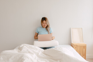 Unhappy asian Thai woman dislike and bored face, using laptop for working at home, putting computer on pillow lap, sitting on white bed, worried trouble at apartment room alone.