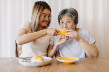 Obraz na płótnie Canvas Happy Asian old woman mother and adult daughter wear glasses, eating croissant bakery and drinking coffee tea at cafe, holding cup and spending happy life time together over white curtain.