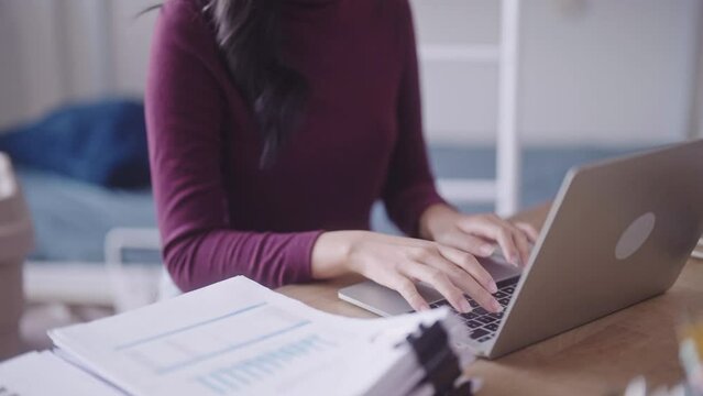 A diligent Asian female student, in casual attire, can be seen completing her homework or typing a research project or thesis on her laptop at her dormitory desk.