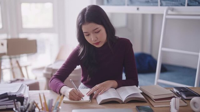 Asian female student, dressed casually, diligently works on her homework or types a research report by referencing study materials at her desk in the dormitory. She is absorbed in her academic tasks.