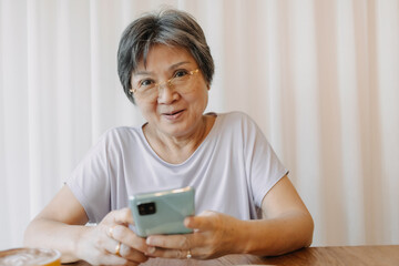 Happy asian old elder woman smiling and looking camera, holding and using phone, chatting or reading news on social, sitting on chair alone over white curtain.