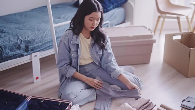 young Asian student in casual clothing is seen unpacking her luggage, taking out clothes and neatly organizing them in her dorm room. She efficiently arranges her belongings in the living space.