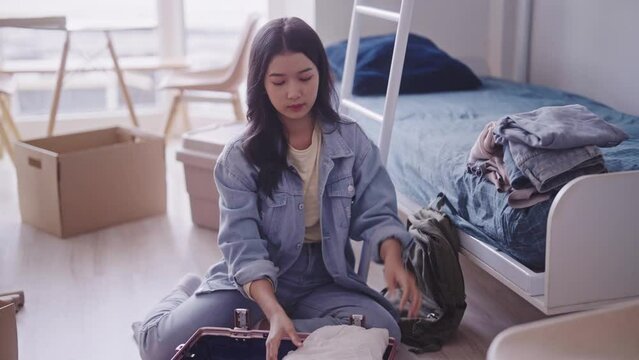 young Asian student in casual clothing is seen unpacking her luggage, taking out clothes and neatly organizing them in her dorm room. She efficiently arranges her belongings in the living space.