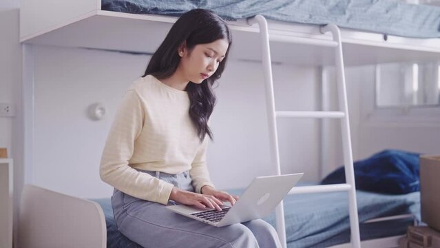 Asian female student, in casual attire, can be seen completing her homework or typing a research project or thesis on her laptop at her dormitory two-tiered bunk bed.