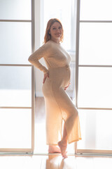 A pregnant woman in a long beige dress stands in profile between the folding doors.