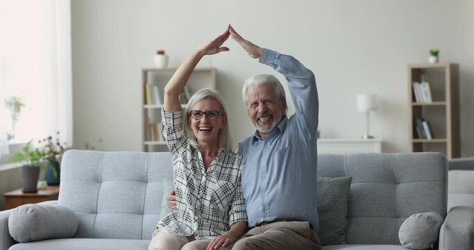 Serious mature elder husband and wife joining hands overhead, showing roof, symbol of protection, insurance, hugging, getting happy, cheerful, laughing, sitting on sofa at home