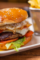 Hamburger with egg and meat, accompanied by french fries, on a plate with creams on a wooden table.