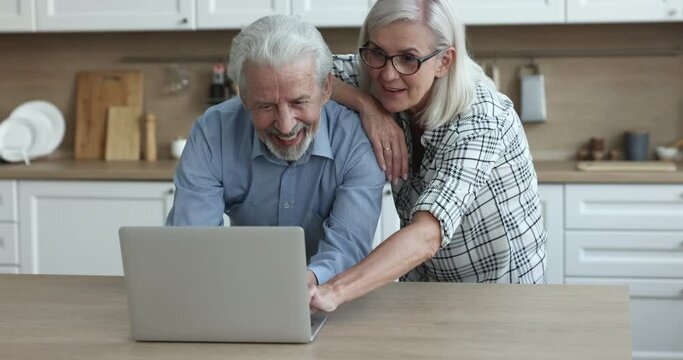 Cheerful older husband and wife using online application, media service, ecommerce app on laptop at home, typing, looking at display, smiling, laughing, enjoying modern Internet technology