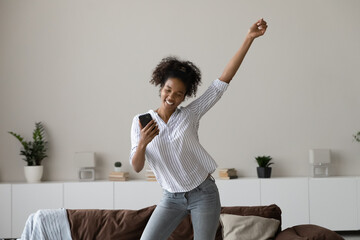 Happy excited millennial Black fit girl dancing at home, holding smartphone, singing to music,...