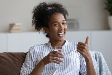 Happy millennial teen African girl drinking fresh water, holding glass, making like thumb up hand...