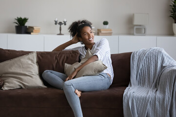 Happy thoughtful young black woman, millennial African girl relaxing on couch in living room, smiling at good thoughts, thinking of future, looking at window away, enjoying leisure at comfortable home