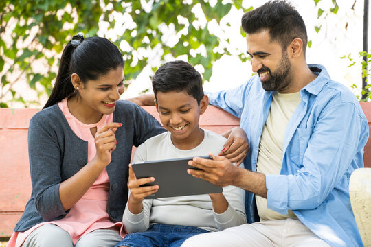 Happy indian couple with kid using digital tablet at park during weekend holidays - concept of technology, cyberspace and modern lifestyle
