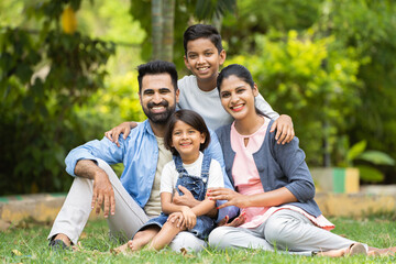 Happy indian couple with kids embracing by looking at camera while sitting at park - concept of relationship, family time and togetherness