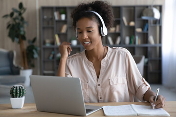 Happy gen Z African student girl in headphones watching webinar on laptop, making video call to...