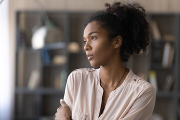 Thoughtful millennial African American girl, student, young woman looking at window away in deep thoughts, thinking over challenges, difficult tasks, feeling bored, sad. Head shot portrait