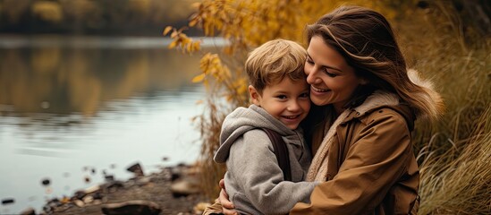 In the autumn season a mother embraces her son against the scenic backdrop of a river - obrazy, fototapety, plakaty
