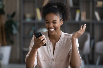 Happy excited young Black woman reading text on mobile phone screen, getting amazing surprising...
