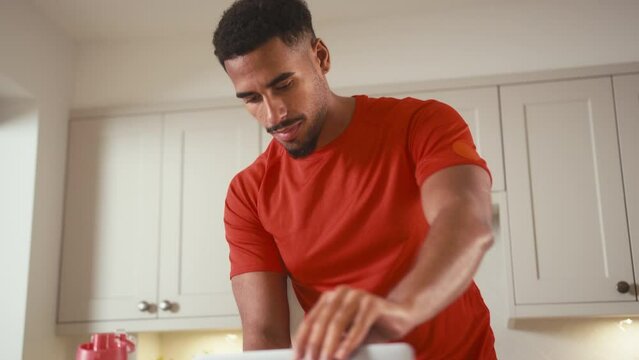 Man wearing fitness clothing with water bottle in kitchen at home working on laptop - shot in slow motion
