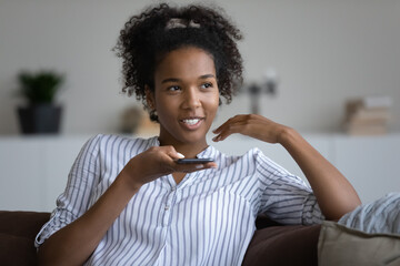 Happy millennial teen Black girl speaking at smartphone speakerphone, recording message for online...