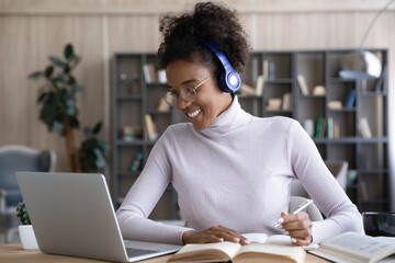 Happy gen Z Black student girl in headphones learning online, watching remote webinar on laptop, writing notes, preparing for exam, doing homework, studying open books on desk. Distance education