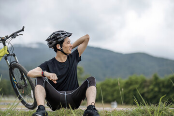 A happy and handsome Asian man in sportswear and a bike helmet enjoys listening to music through his earbuds while riding a bike on the country roads on the weekend.
