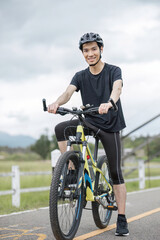 A happy and handsome Asian man in sportswear and a bike helmet, enjoys listening to music through his earbuds while riding a bike on the country roads on the weekend.