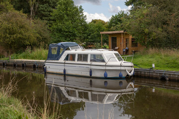 A narrow boat is moored on a canal in front of a wooden shed. It is perfectly reflected in the still water - 672570031