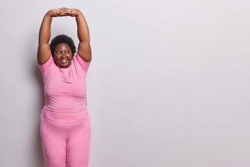 Sporty lifestyle. Indoor shot of young happy smiling plump darkskinned woman standing on left isolated on white background in pink tracksuit doing stretching exercise with blank space for promotion - obrazy, fototapety, plakaty