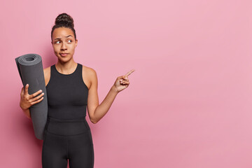 Horizontal shot of Latin woman with dark hair gathered in bun poses with fitness mat under arm dressed in black tracksuit isolated over pink background. People yoga and fitness training concept