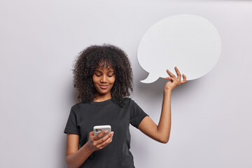 Horizontal shot of pretty curly haired African woman holds modern mobile phone and blank speech bubble for your advertising content dressed in casual black t shirt isolated over white background.