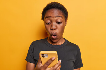 Photo of stunned dark skinned woman has astonished expression as she gazes at her smartphone screen...