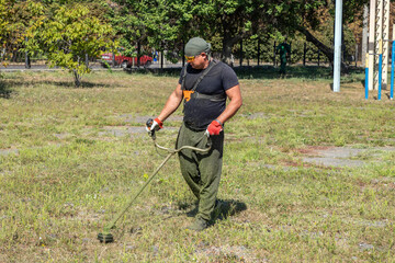 Man gardener mowing ragweed sprouts or plants, ambrosia artemisiifolia that causing allergy summer and autumn. Man mows grass with a gasoline or electric scythe, or petrol lawn trimmer in public park.