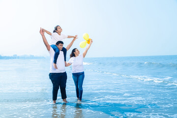 Happy young indian family of three wearing white t-shirt and blue jeans walking at beach, .Father carrying his daughter on shoulders, enjoying summer vacations. Travel and holidays. Copy space.