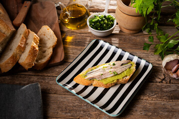 Spanish tapa of a slice of bread spread with guacamole and some anchovies in vinegar on top, on a rustic table.