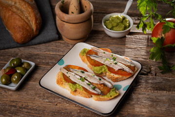 Spanish tapa with a slice of bread spread with guacamole, a sliced tomato and some anchovies in vinegar on top, on a rustic table.