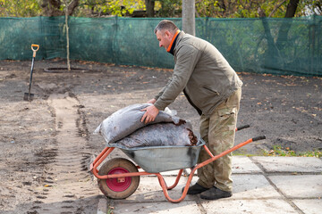 A professional gardener, middle-aged male worker with wheelbarrow full of bags with fertilazer or...