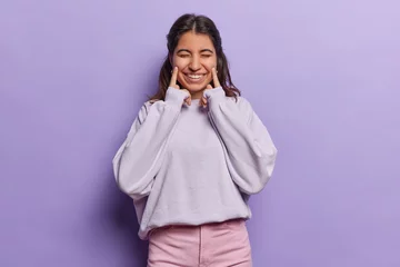 Fotobehang Beautiful Iranian woman forces happy smile on face keeps fingers on cheeks keeps eyes closed tries to be happy has eyes closed dressed in casual sweatshirt and pink trousers isolated over purple wall © Wayhome Studio
