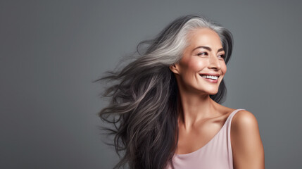 Sensual middle-aged beautiful Asian woman with long grey hair, her eyes closed, body and face care concept. Charming mature lady stands in profile isolated on grey studio background - obrazy, fototapety, plakaty