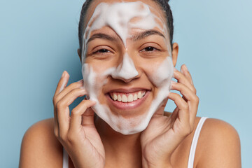 Close up shot of cheerful woman washing face with foam cleanser smiles gladfully shows white teeth...